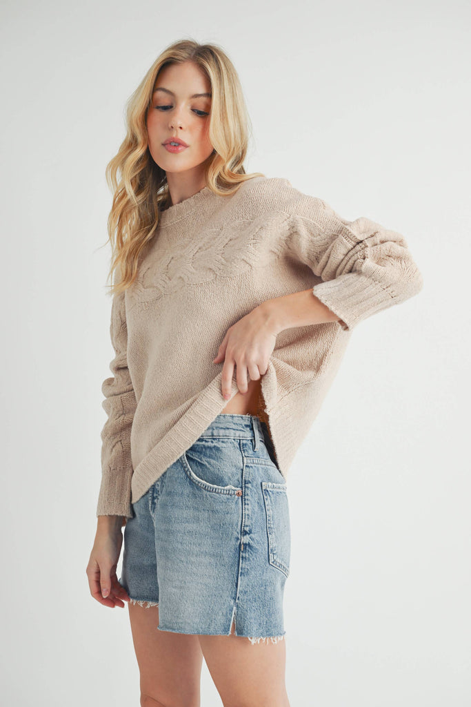 Woman wearing a beige cable-knit sweater with relaxed fit and cable details on the chest and sleeves.