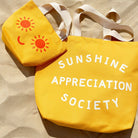 Yellow canvas tote bag with “Sunshine Appreciation Society” printed in white, held up by a hand in front of a sage green wall