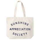 Cream canvas tote bag with “Sunshine Appreciation Society” printed in navy, filled with flip-flops, sunglasses, and travel essentials on a pink background