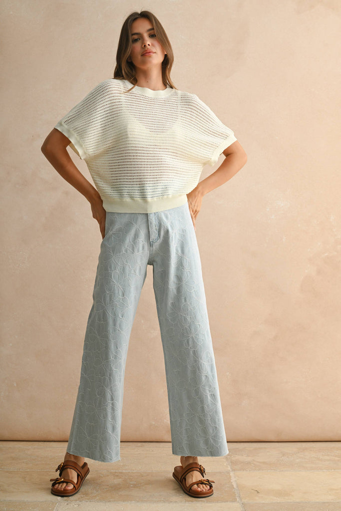 Model wearing a cream open-knit short sleeve sweater with open back detail, paired with embroidered light-wash denim pants.