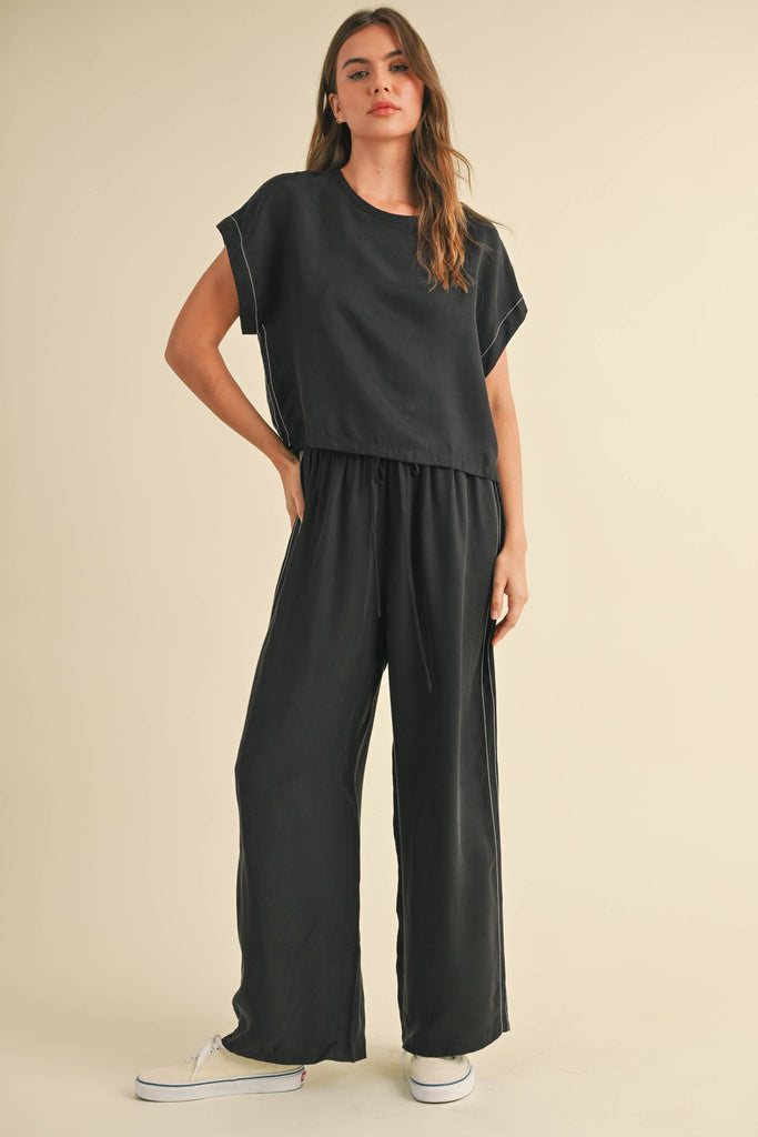 Model wearing a black two-piece lounge set with white contrast stitching, featuring a cap-sleeve top and drawstring wide-leg pants.