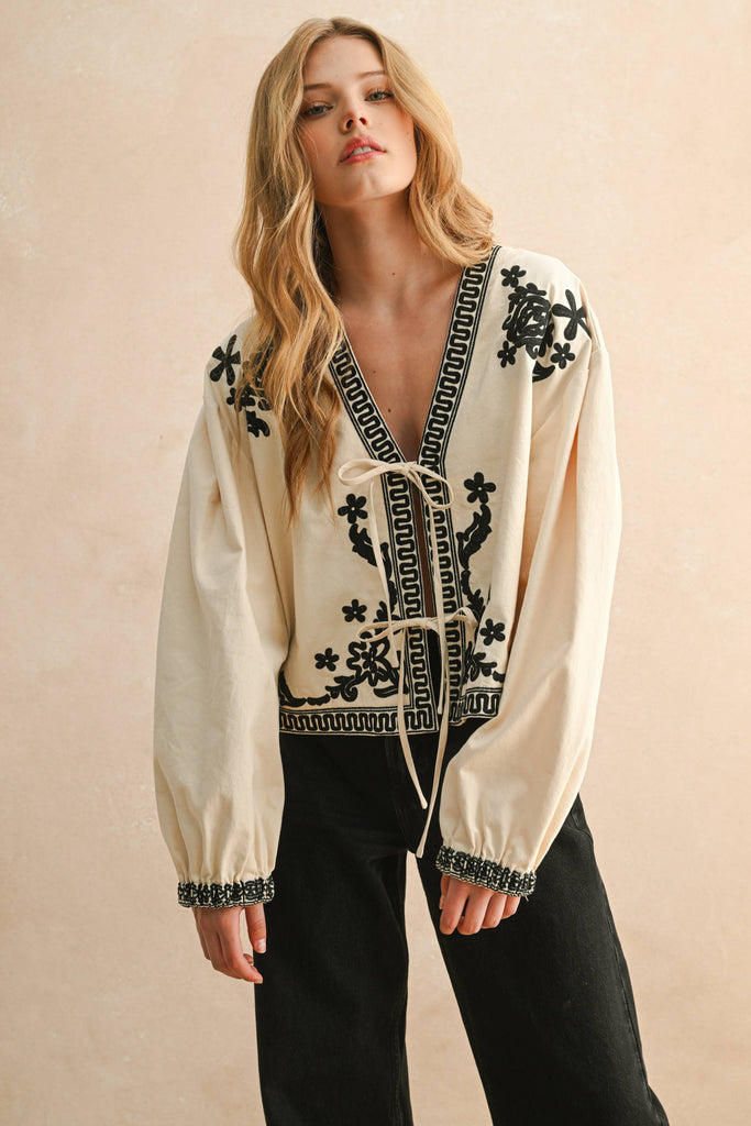 Woman wearing cream blouse with black floral embroidery, lace-up front, and long billowy sleeves.