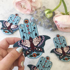 Large embroidered patch displaying a Hamsa integrated with butterfly, evil eyes, crescent moons, arrows, and floral motifs in a vibrant boho color palette.