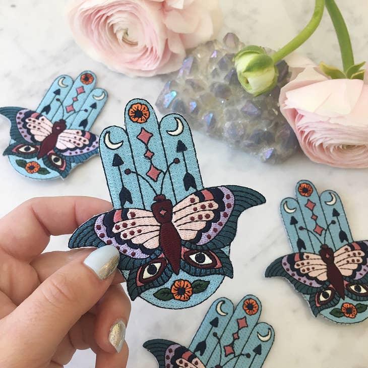 Large embroidered patch displaying a Hamsa integrated with butterfly, evil eyes, crescent moons, arrows, and floral motifs in a vibrant boho color palette.
