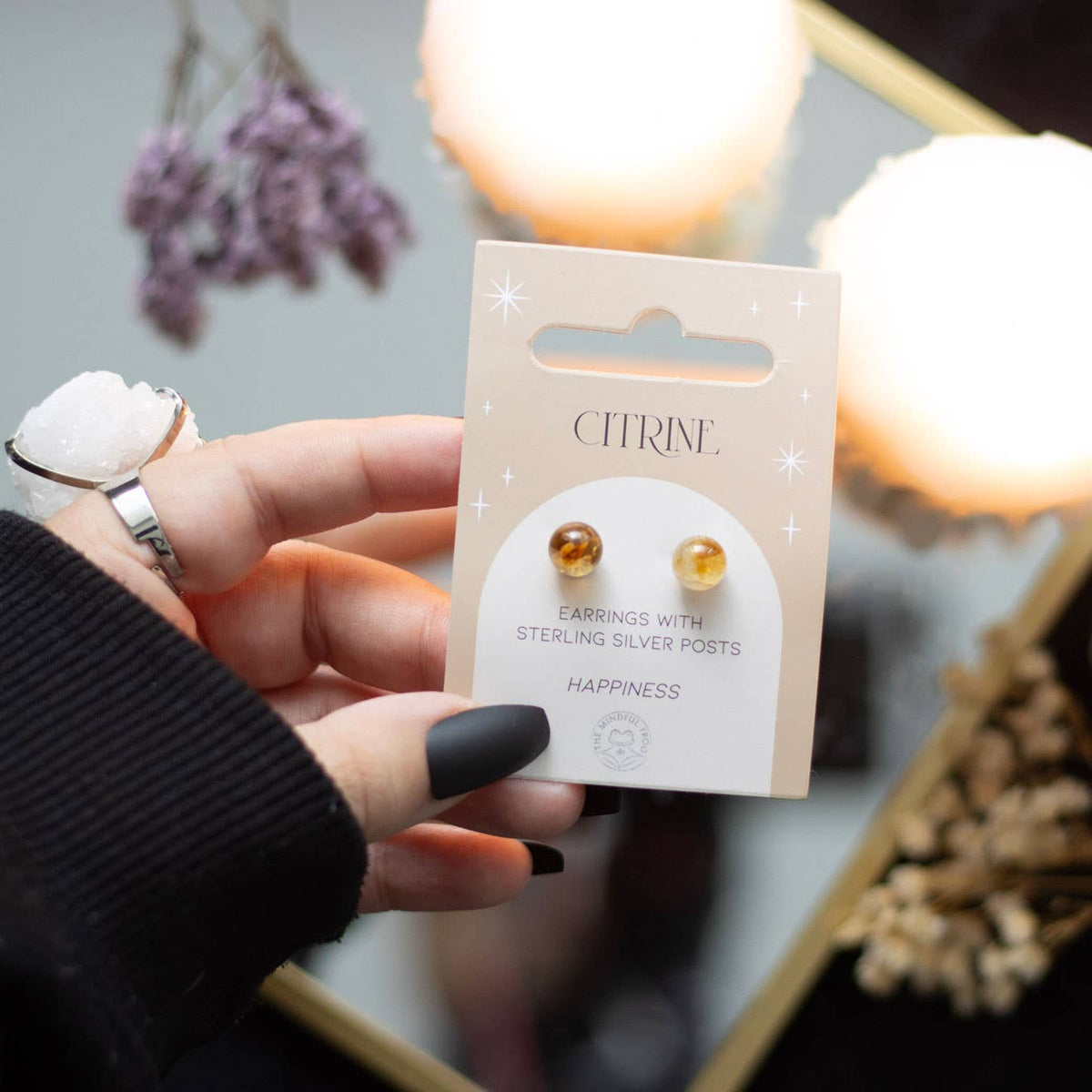 A pair of citrine crystal stud earrings displayed on a matching backing card, held in a hand with a serene background.