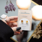 A pair of citrine crystal stud earrings displayed on a matching backing card, held in a hand with a serene background.