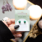 Hand holding a pair of malachite crystal stud earrings on a matching backing card with the text “Promotes Confidence.”