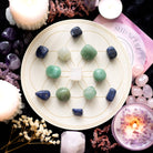 Seed of Life Crystal Grid Set featuring 6 sodalite tumblestones, 5 aventurine tumblestones, 1 clear quartz tumblestone, and an engraved wooden grid, surrounded by candles and decorative flowers.