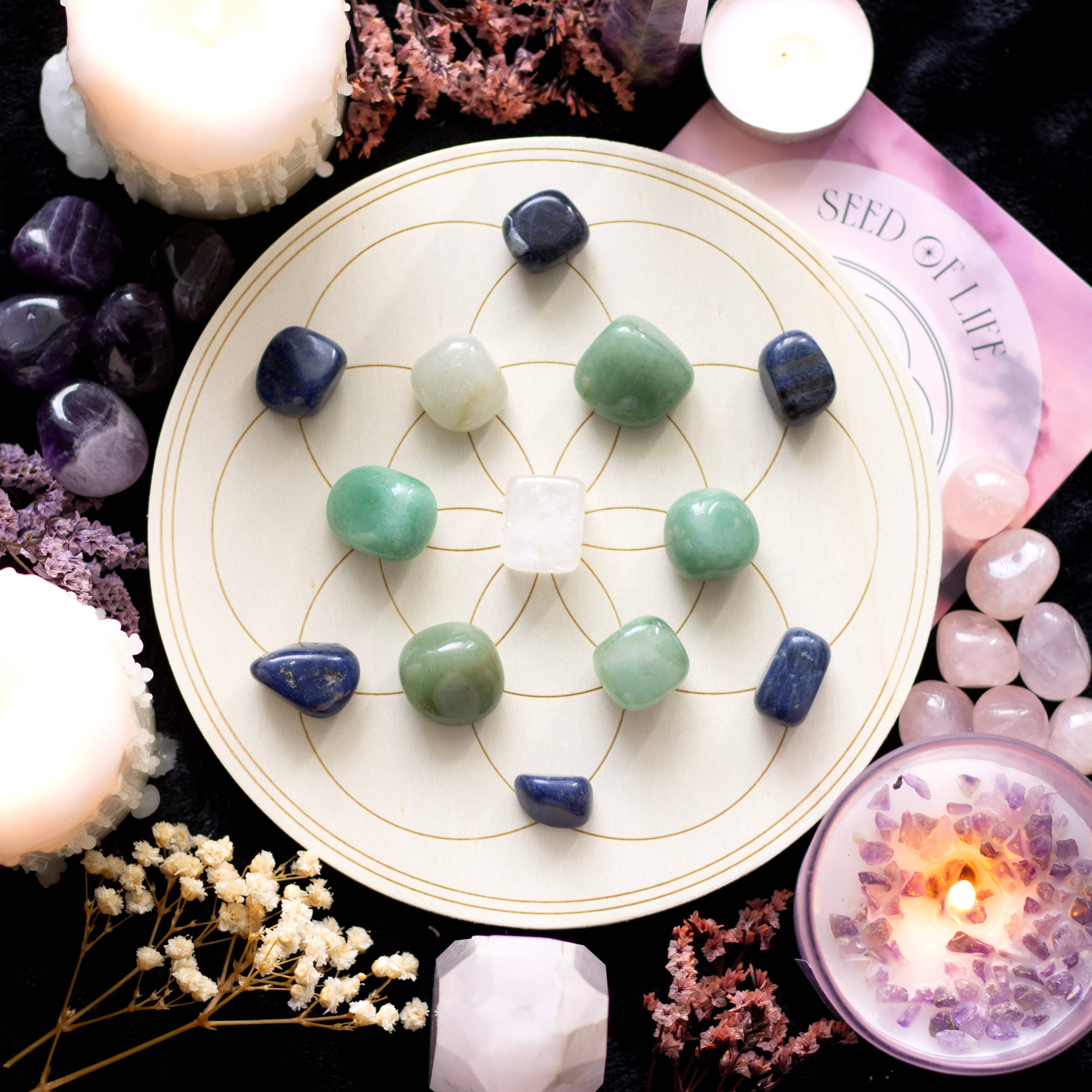 Seed of Life Crystal Grid Set featuring 6 sodalite tumblestones, 5 aventurine tumblestones, 1 clear quartz tumblestone, and an engraved wooden grid, surrounded by candles and decorative flowers.